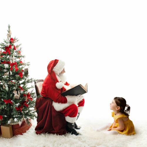santa sitting with a christmas tree reading to little girl