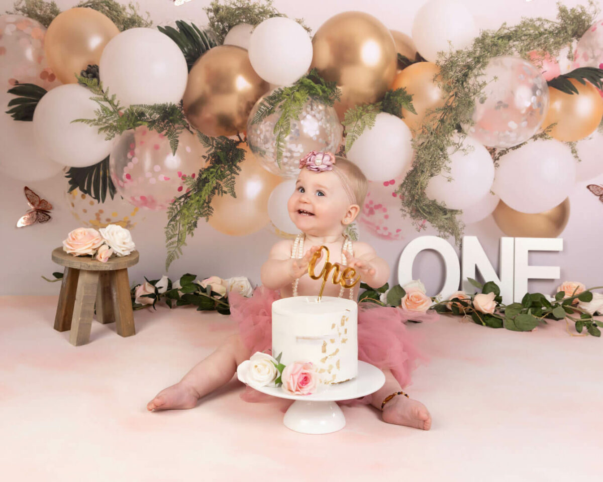 happy baby in pink tutu with pink and gold balloons and birthday cake