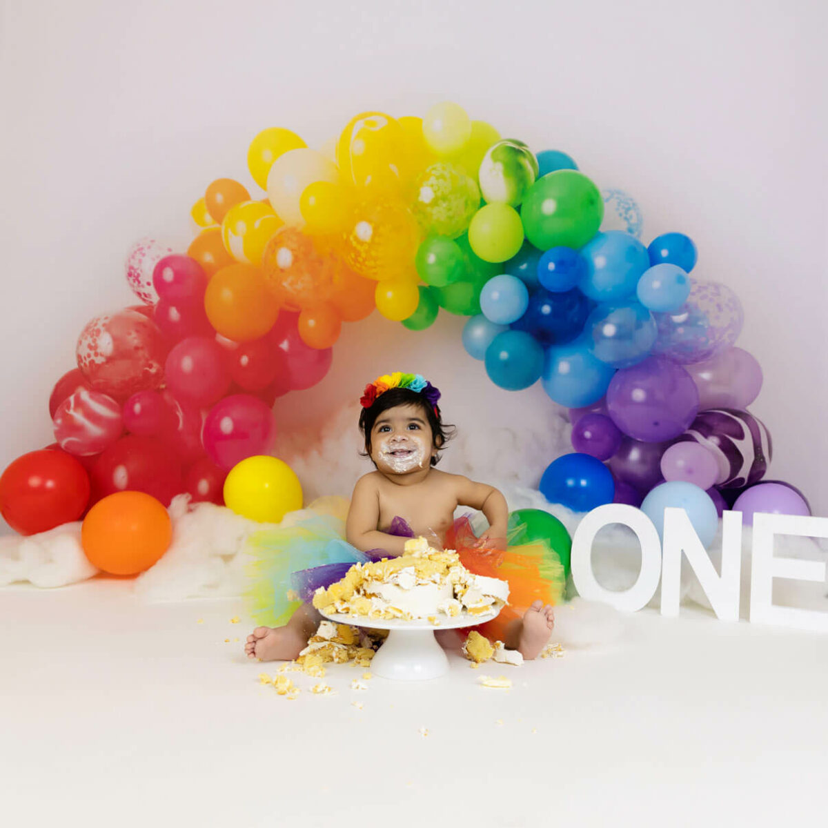 baby with rainbow balloons trying birthday cake