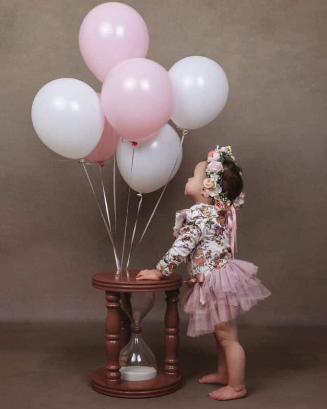baby in pink with pink flower crown looking at pink and white balloons on hourglass