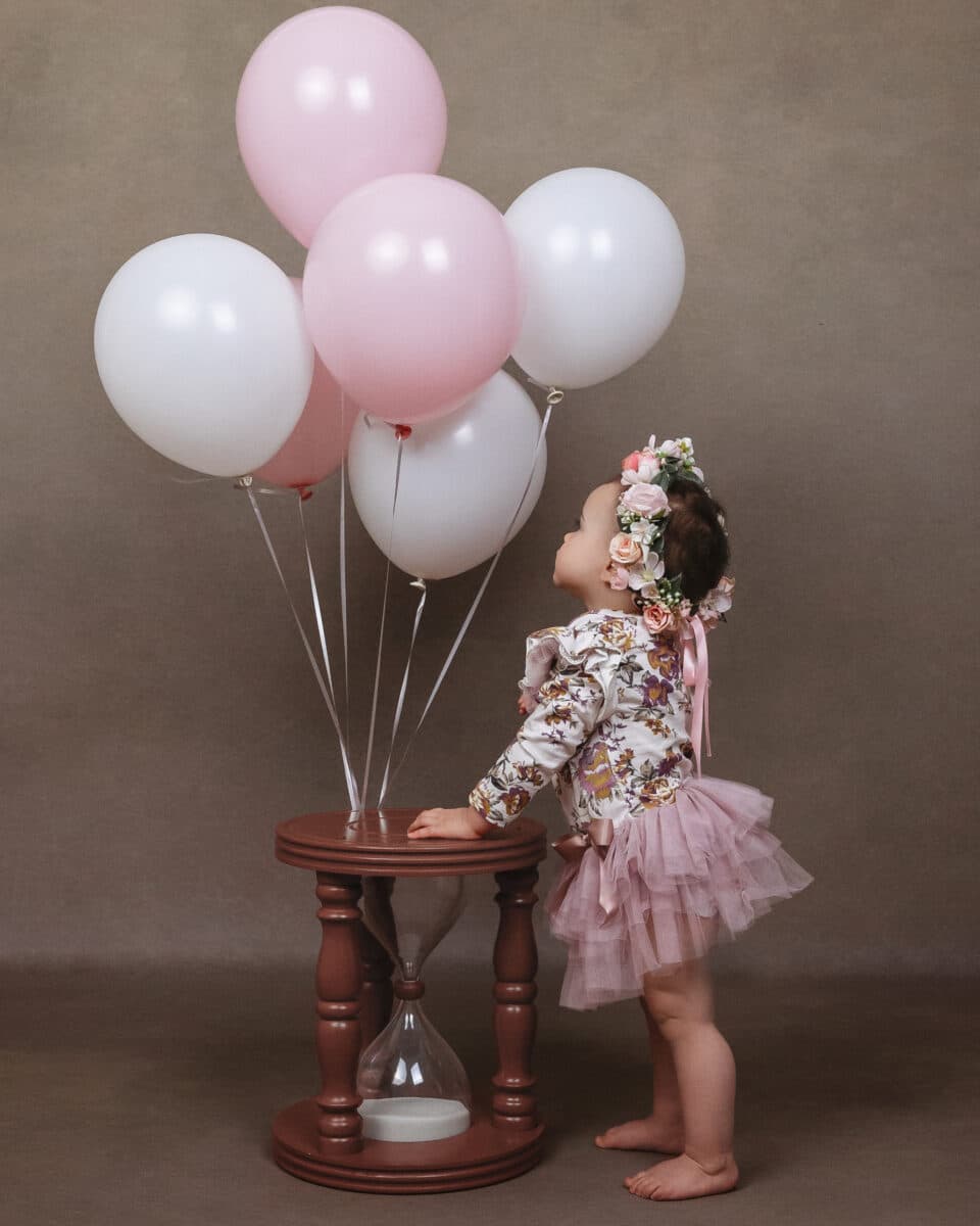 baby in pink with pink flower crown looking at pink and white balloons on hourglass