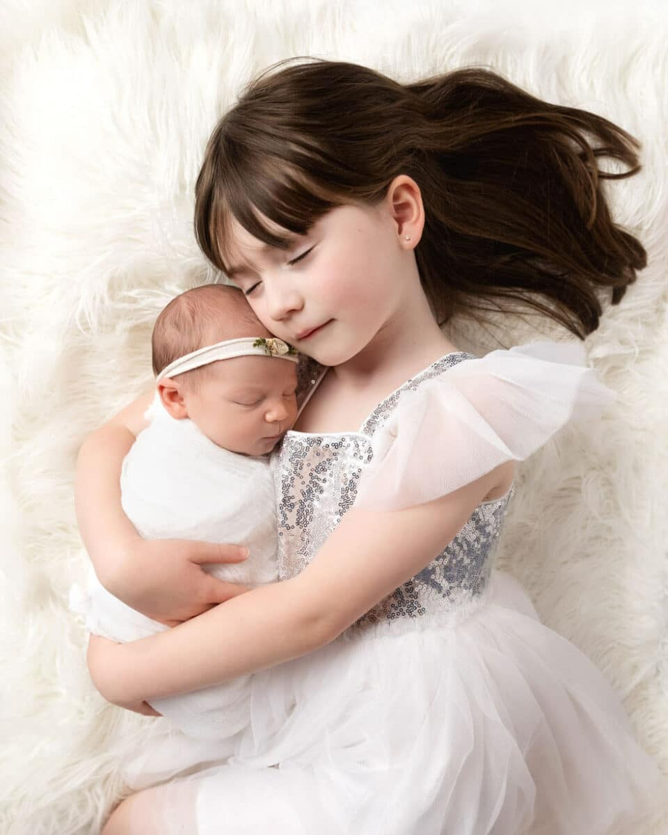 sister cuddling newborn baby on white plush rug